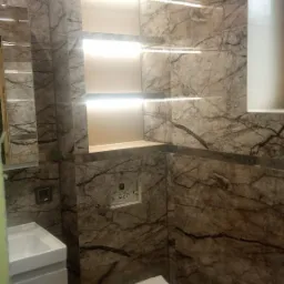 Wykończona łazienka w Kazuniu Bielanach z marmurowymi płytkami, podwieszaną toaletą i umywalką oraz oświetlonymi półkami w ścianie.