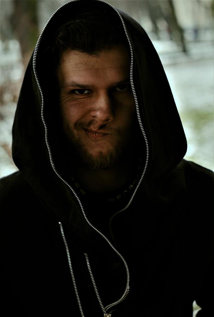 Portret mężczyzny w czarnej bluzie z kapturem, z lekkim uśmiechem i spojrzeniem w dół, na rozmytym tle zewnętrznym.