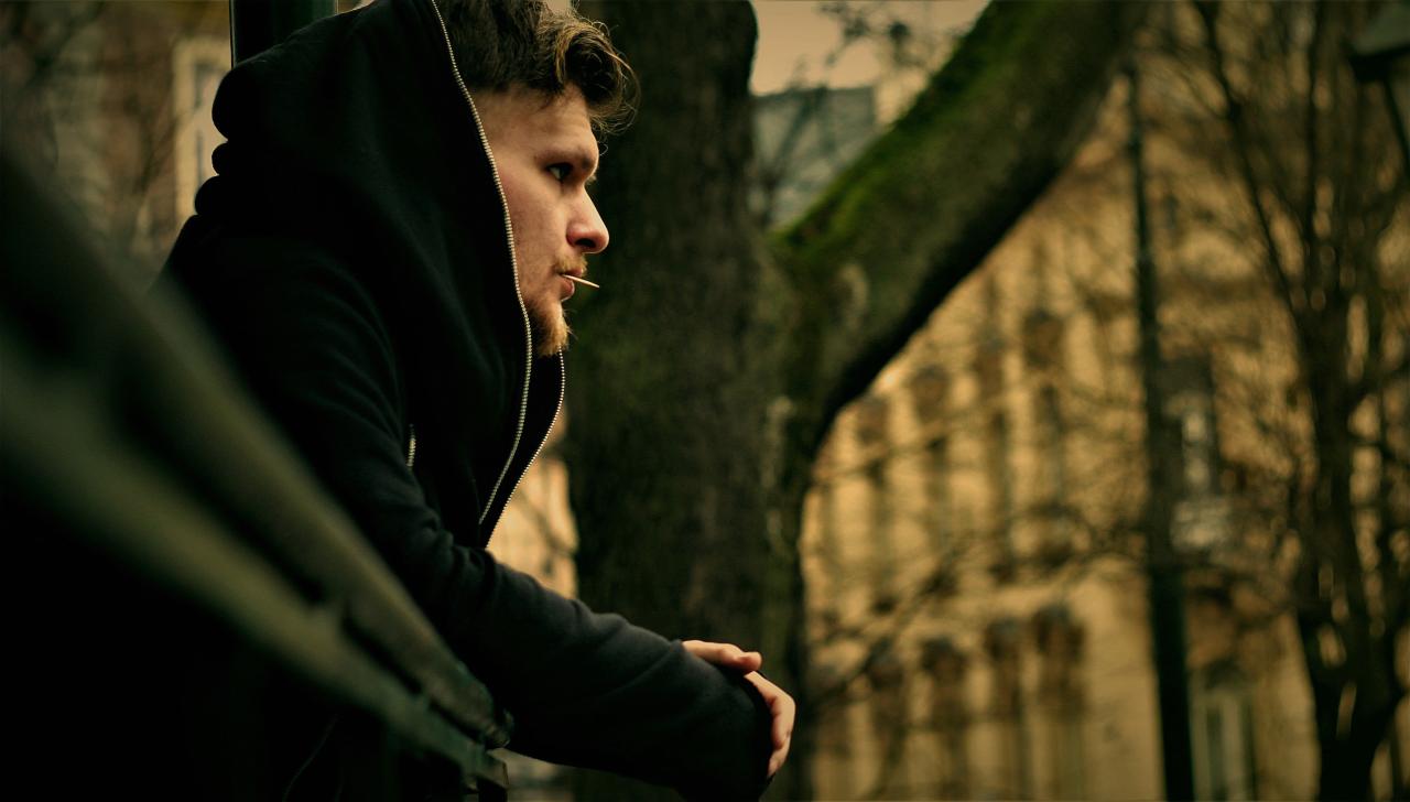 Mężczyzna w czarnej bluzie z kapturem, z zamyślonym wyrazem twarzy i wykałaczką w ustach, oparty o metalową barierkę na tle drzewa i budynku.