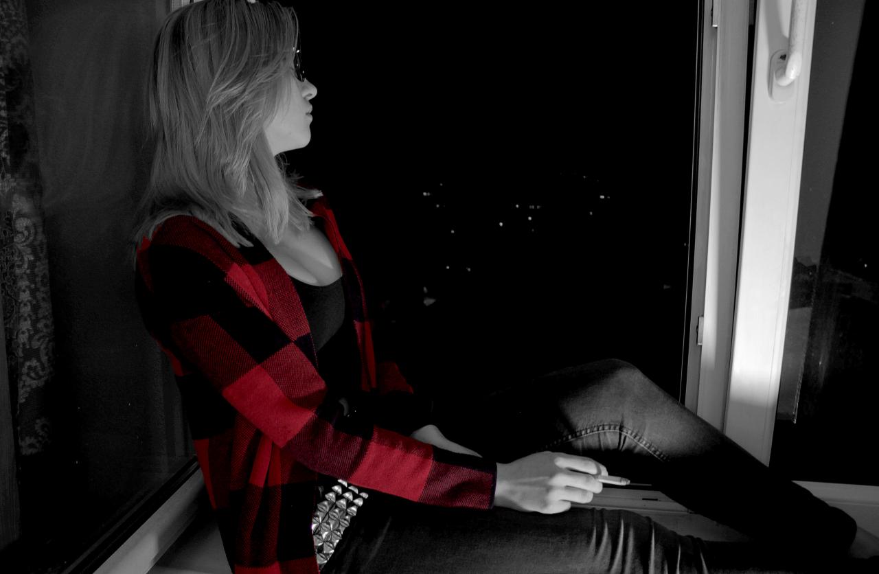 Kobieta w czerwono-czarnej koszuli w kratę siedzi na parapecie okna, patrząc na nocne miasto, częściowo w czerni i bieli.