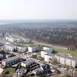 Panoramiczny widok z lotu ptaka na osiedle bloków mieszkalnych sąsiadujące z lasem i jeziorem w oddali, uchwycony w pochmurny dzień.