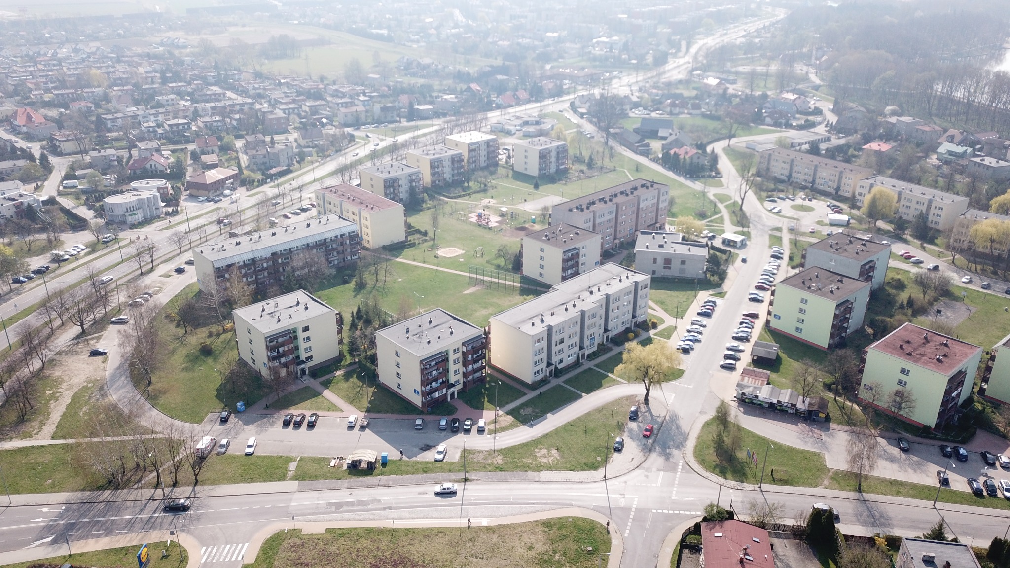 Widok z lotu ptaka na osiedle bloków z placem zabaw, parkingami i zielenią w słoneczny dzień.