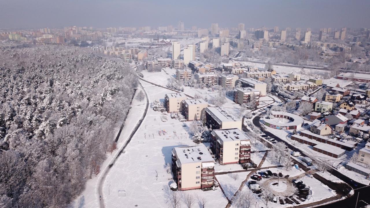 Zimowy krajobraz miejski z lotu ptaka: ośnieżony las graniczy z blokami mieszkalnymi, w tle panorama miasta pod lekką mgłą.
