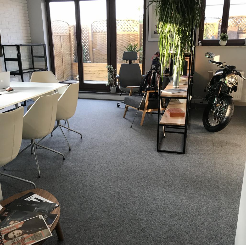 Jasne biuro z białym stołem, krzesłami i motocyklem zaparkowanym w rogu, widoczne rośliny doniczkowe i regał.