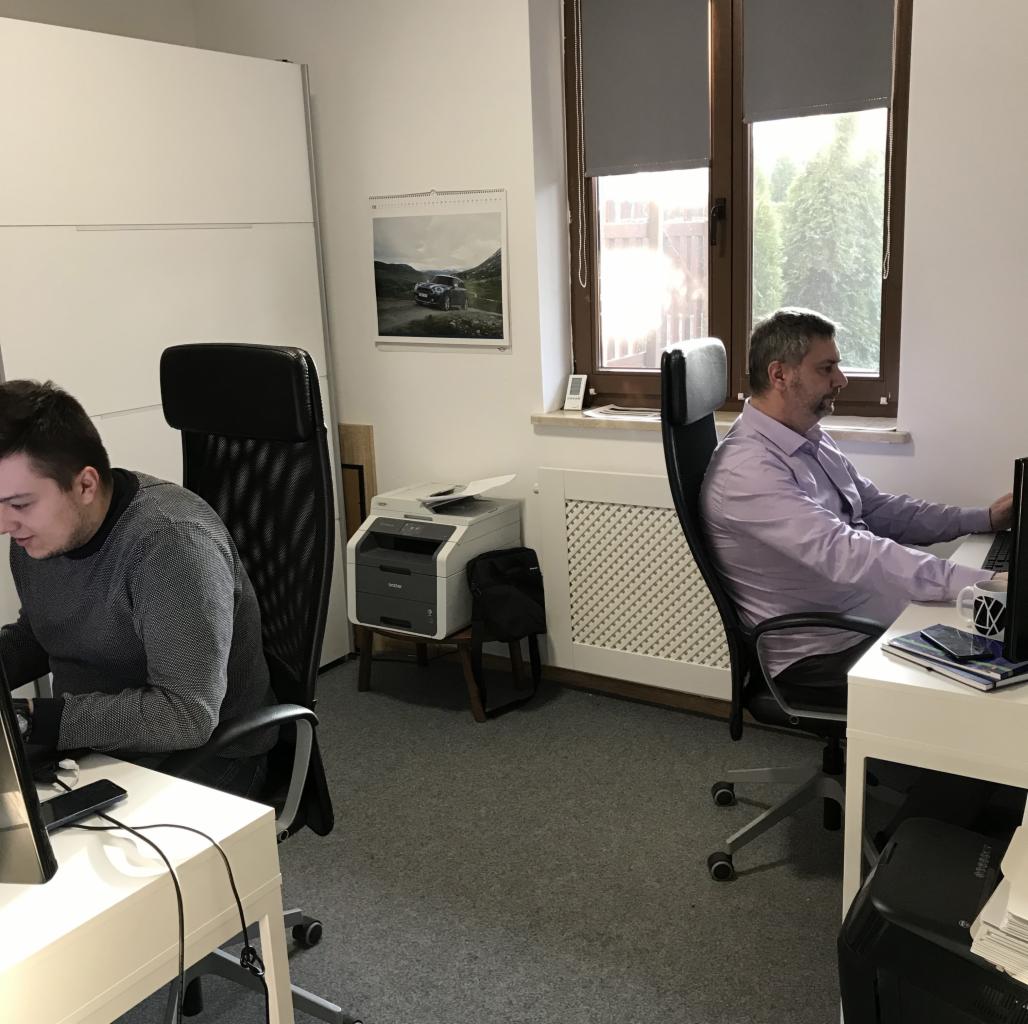 Dwaj mężczyźni pracują przy biurkach w biurze, jeden w szarej bluzie, drugi w fioletowej koszuli, z widocznym kalendarzem z samochodem na ścianie i drukarką w tle.