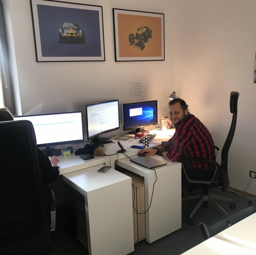 Biuro z dwoma stanowiskami pracy, na ścianie wiszą plakaty z samochodami, pracownik w czerwonej koszuli uśmiecha się do kamery.