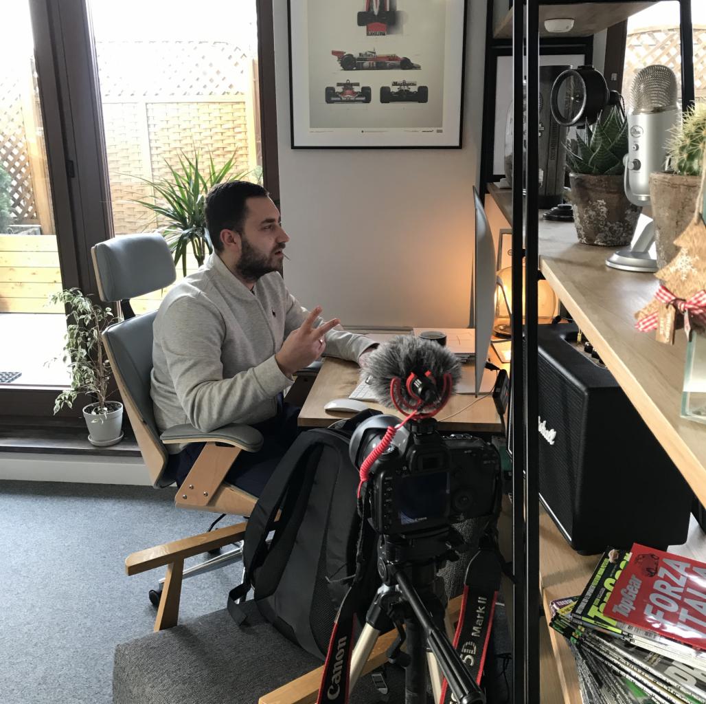 Mężczyzna siedzący przy biurku z mikrofonem i kamerą, prawdopodobnie nagrywający wideo lub prowadzący transmisję na żywo, w tle plakat z bolidami Formuły 1