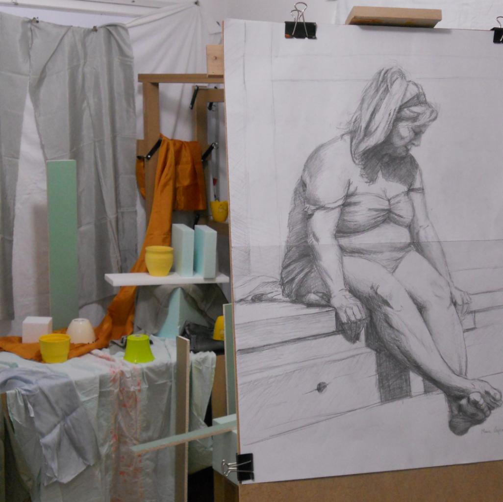 Szkic ołówkiem przedstawiający siedzącą kobietę, ustawiony w pracowni artystycznej z rekwizytami i draperiami, w tle widoczne geometryczne bryły i kolorowe naczynia.