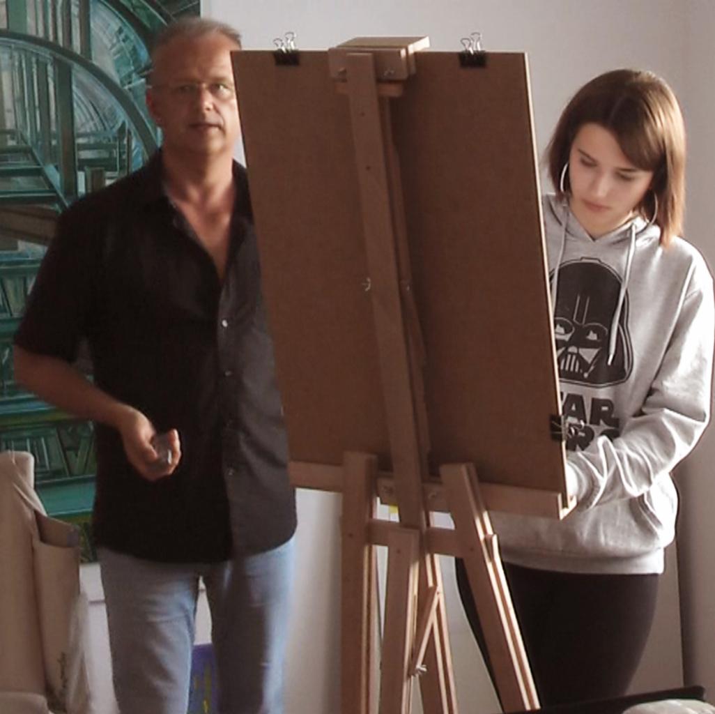 Instruktor prowadzi zajęcia z rysunku, demonstrując technikę obok uczennicy pracującej przy sztaludze.