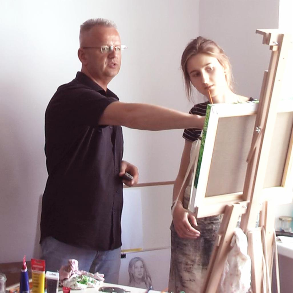 Instruktor malarstwa demonstruje technikę na płótnie uczennicy, w tle widoczny szkic portretu.