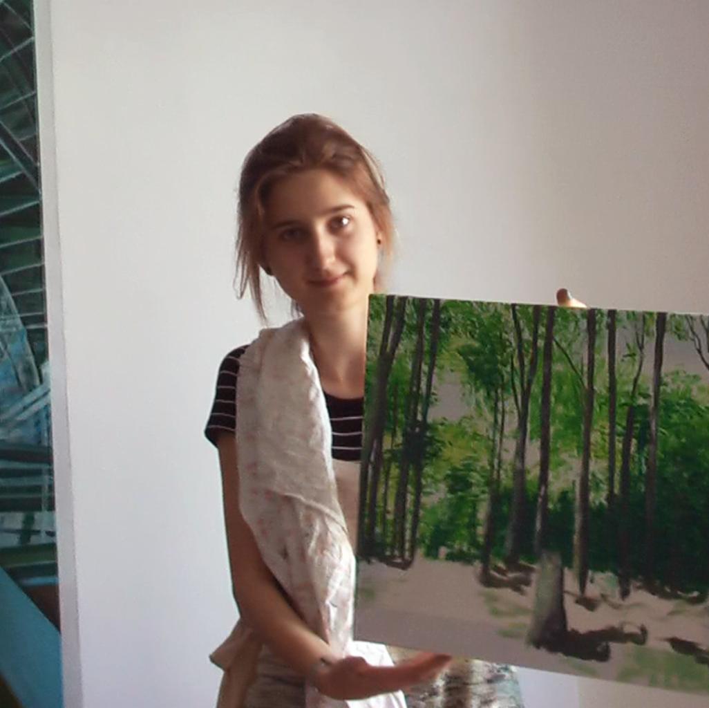 Uczestniczka kursu malarstwa prezentuje obraz lasu z ciemnymi pniami drzew i zielonymi liśćmi, trzymając go przed sobą w jasnym pomieszczeniu.