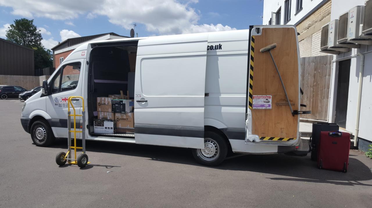 Biały van z otwartymi drzwiami bocznymi i tylnymi, załadowany kartonami i z wózkiem transportowym obok, sugerujący proces załadunku lub rozładunku.