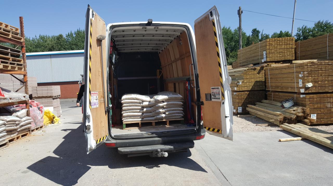 Otwarty tył białego vana z paletą worków w środku, widoczne drewniane deski i palety w tle.