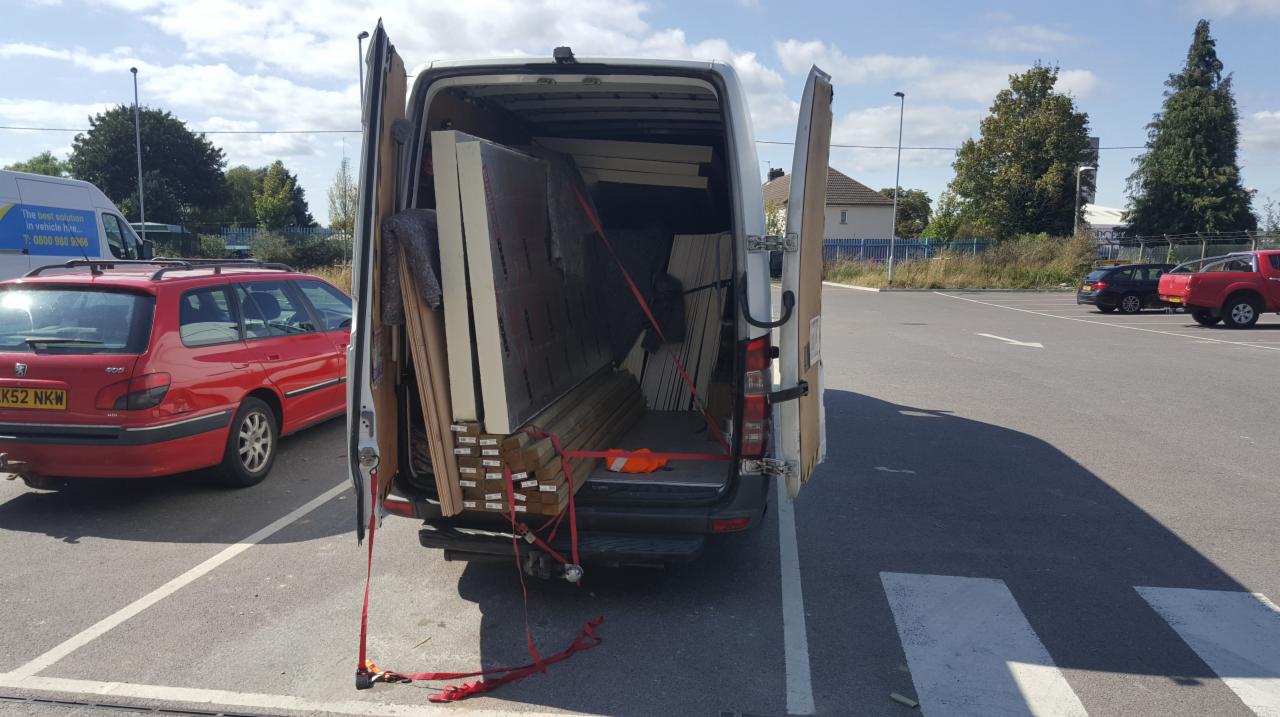 Załadunek białego busa z otwartymi drzwiami: płyty meblowe, deski i belki zabezpieczone czerwonymi pasami transportowymi na parkingu.