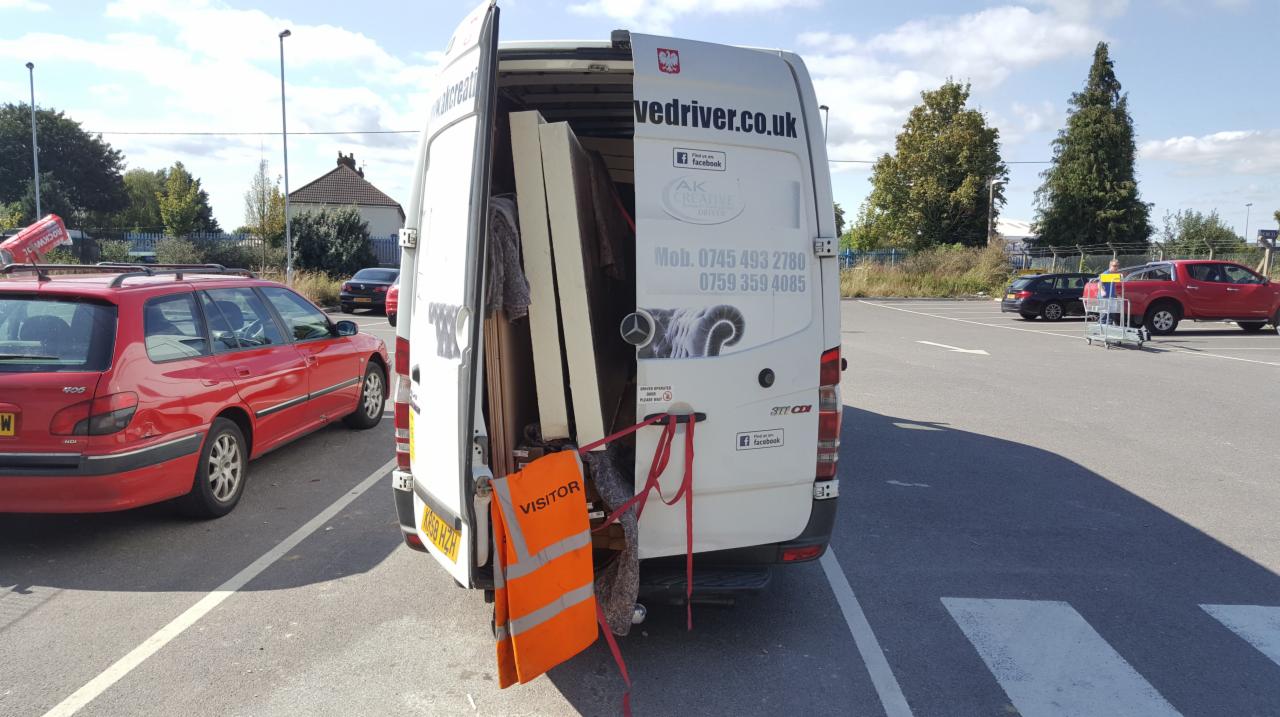 Otwarty tył białego busa transportowego, wypełnionego meblami zabezpieczonymi do transportu, z wiszącą kamizelką odblaskową 'VISITOR', zaparkowanego na parkingu.