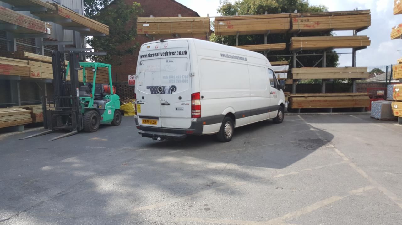 Biały bus dostawczy z logo firmy zaparkowany na placu budowlanym obok zielonego wózka widłowego i składowanego drewna.