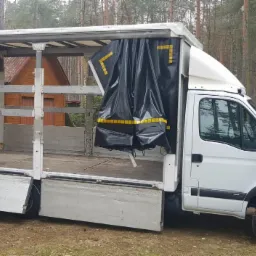 Biały samochód dostawczy z otwartą przestrzenią ładunkową i rozpiętą czarną plandeką z żółtymi oznaczeniami, zaparkowany na trawiastym terenie z widocznym lasem w tle.