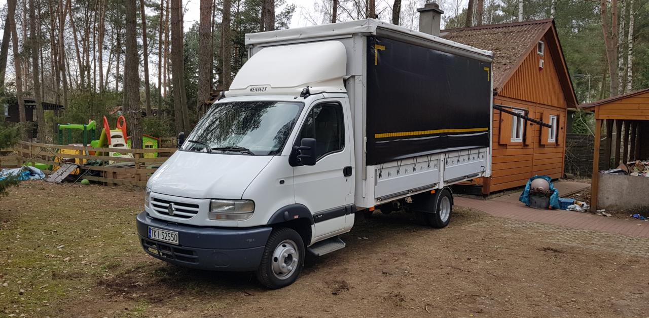Biały samochód dostawczy Renault z plandeką na posesji w Stanisławowie Pierwszym, widoczny fragment drewnianego domu i placu zabaw w tle.