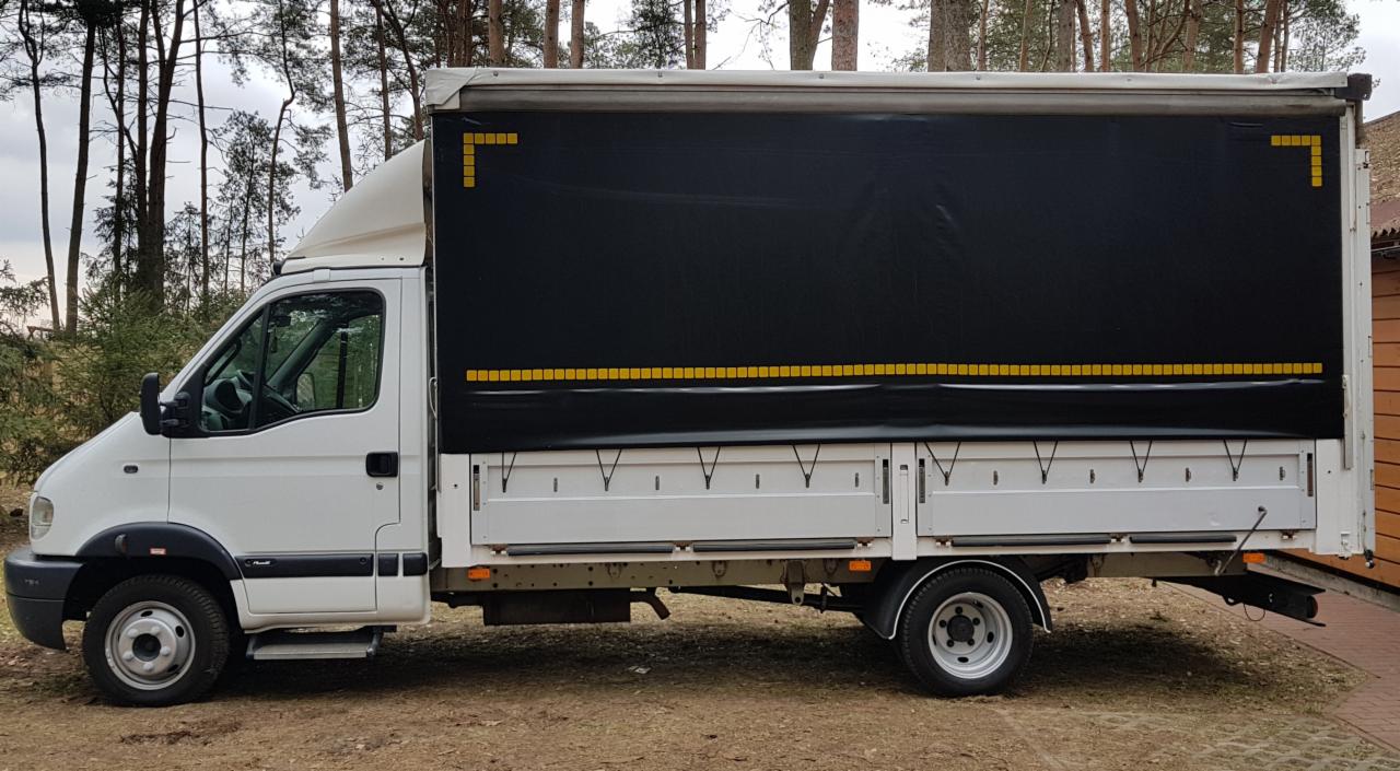 Biały samochód dostawczy z czarną plandeką ozdobioną żółtymi elementami, zaparkowany na ziemi z widocznym lasem w tle.