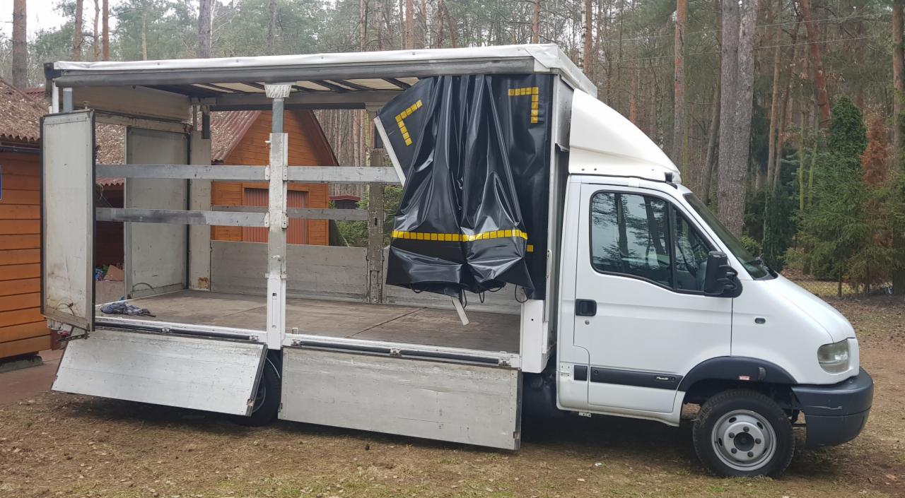 Biały samochód dostawczy z otwartą przestrzenią ładunkową i rozpiętą czarną plandeką z żółtymi oznaczeniami, zaparkowany na trawiastym terenie z widocznym lasem w tle.