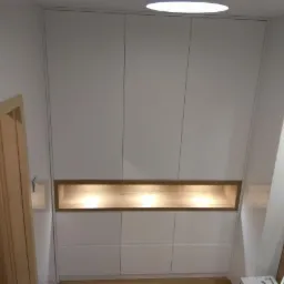 Biała zabudowa meblowa od podłogi do sufitu z podświetlaną drewnianą wnęką, widok z dołu, obok wejście do pomieszczenia.