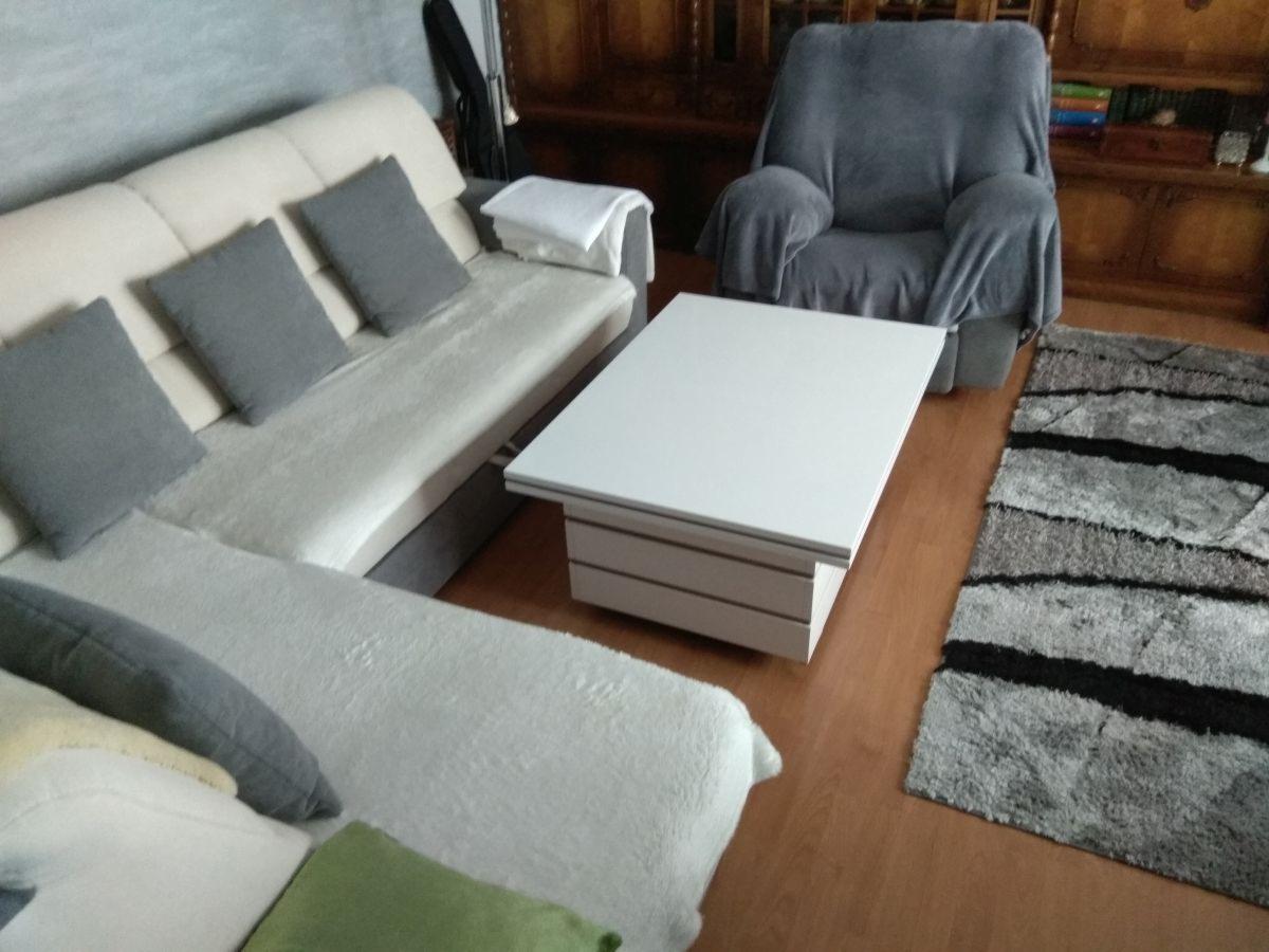 Jasna, narożna sofa z szarymi poduszkami, biały stolik kawowy z szufladami, szary fotel z narzutą i dywan w paski na drewnianej podłodze.