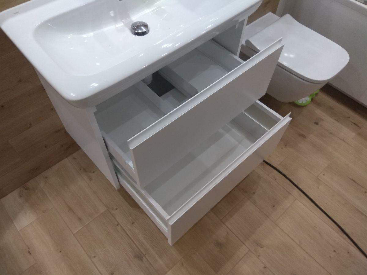 Biała umywalka z otwartymi szufladami w łazience z drewnianą podłogą, widoczny fragment toalety w tle.