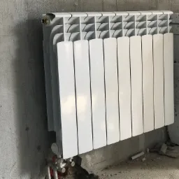 Biały, aluminiowy grzejnik panelowy zamontowany na surowej, betonowej ścianie w nowym budynku, widoczne rury przyłączeniowe i stos płyt betonowych w tle.