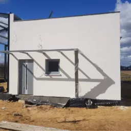 Surowy stan domu murowanego z szarą stolarką okienną i drzwiową, widoczne niedociągnięcia w elewacji, fundamenty zabezpieczone folią, stalowa konstrukcja w tle.