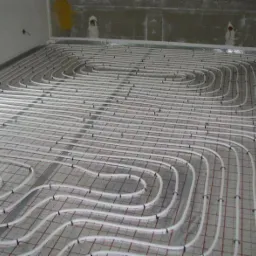 Białe rurki ogrzewania podłogowego rozłożone na siatce z czerwonymi liniami, przygotowane do zalania wylewką w surowym pomieszczeniu.