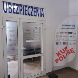 Wejście do agencji ubezpieczeniowej z napisem 'UBEZPIECZENIA' nad drzwiami, szyldem 'NOWATOR Agencja Ubezpieczeń' i listą firm ubezpieczeniowych na fladze obok, oraz informacjami na szybie...