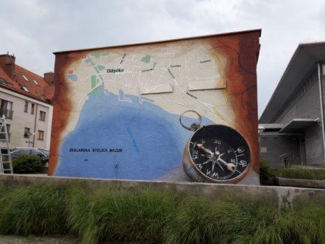 Mural przedstawiający mapę Giżycka z kompasem, umieszczony na ścianie budynku w plenerze.