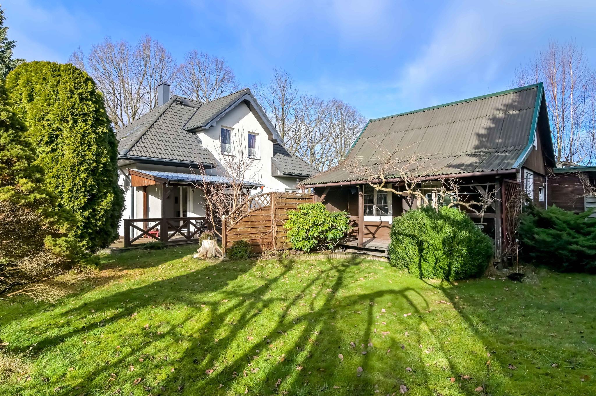 Kopalino: Dwa domy z ogrodami, zróżnicowana architektura, zielony trawnik i drzewa bez liści w tle, jasne niebo. Unikalne cienie drzew na trawie.