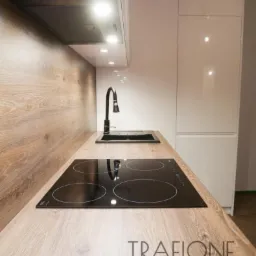 Kuchenny blat z drewnianym wzorem, czarna płyta indukcyjna i zlew, minimalistyczna bateria, białe szafki wiszące i okap, logo firmy na blacie.