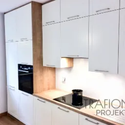 Jasna kuchnia z białymi szafkami, drewnianym blatem i zabudowaną piekarnikiem, z czarną płytą indukcyjną i garnkiem na blacie.