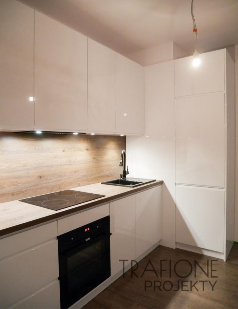 Nowoczesna kuchnia z białymi, lakierowanymi frontami, drewnianym blatem i panelem ściennym, czarnym zlewem i płytą indukcyjną; widoczne logo firmy na dolnej szafce.