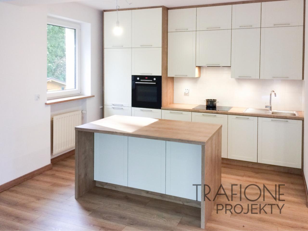 Jasna kuchnia z wyspą, drewniany blat i obudowa, białe fronty szafek, zabudowa kuchenna pod sufit, widoczne oświetlenie podszafkowe i płyta indukcyjna z garnkiem.