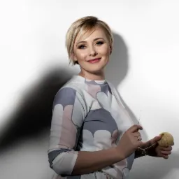 Portret uśmiechniętej kobiety z blond włosami, trzymającej w dłoniach kłębek wełny i szydełko na tle białej ściany z cieniem.