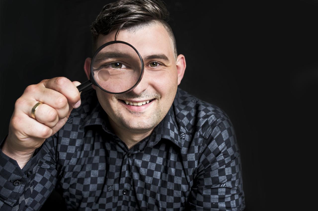 Uśmiechnięty mężczyzna w koszuli w kratę, z obrączką na palcu, patrzy przez szkło powiększające na tle czarnego tła.