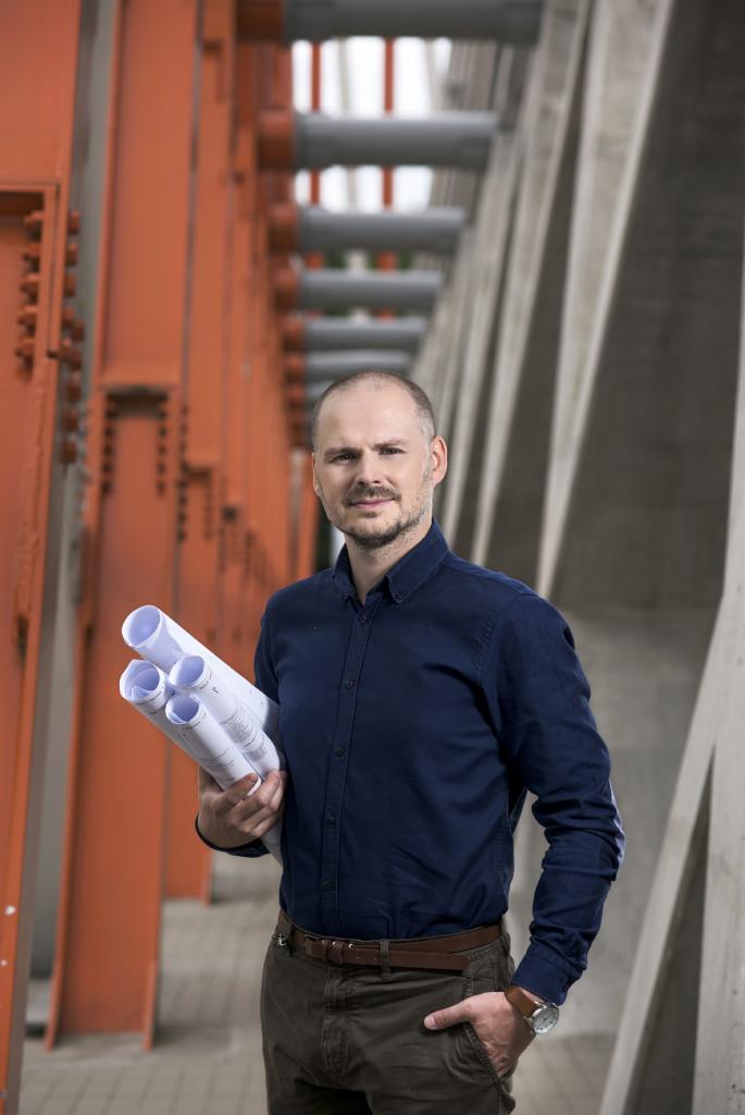 Mężczyzna w niebieskiej koszuli trzymający zwitki planów na tle konstrukcji stalowej i betonowej.