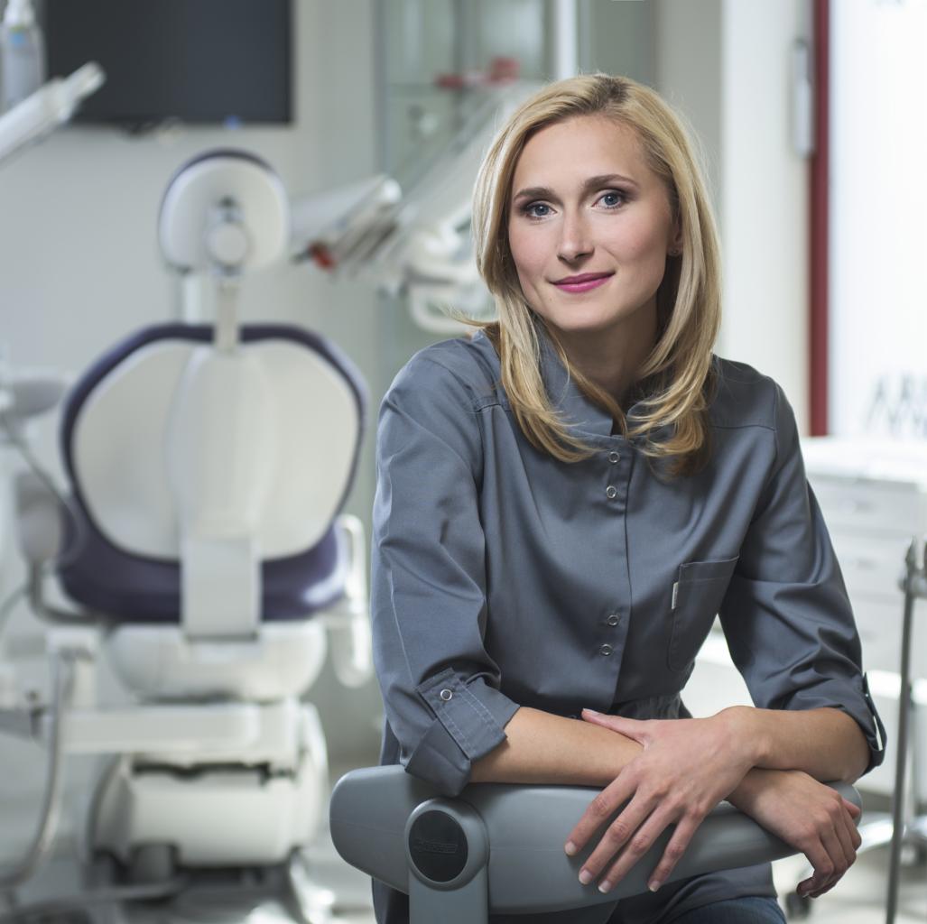 Portret uśmiechniętej blondynki w szarym uniformie medycznym, opierającej się o fotel dentystyczny w nowoczesnym gabinecie.