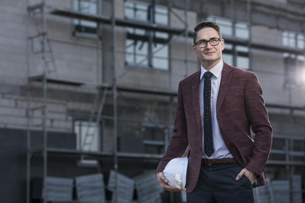 Młody mężczyzna w garniturze i okularach, trzymający biały kask na tle budynku w trakcie budowy ze rusztowaniami.
