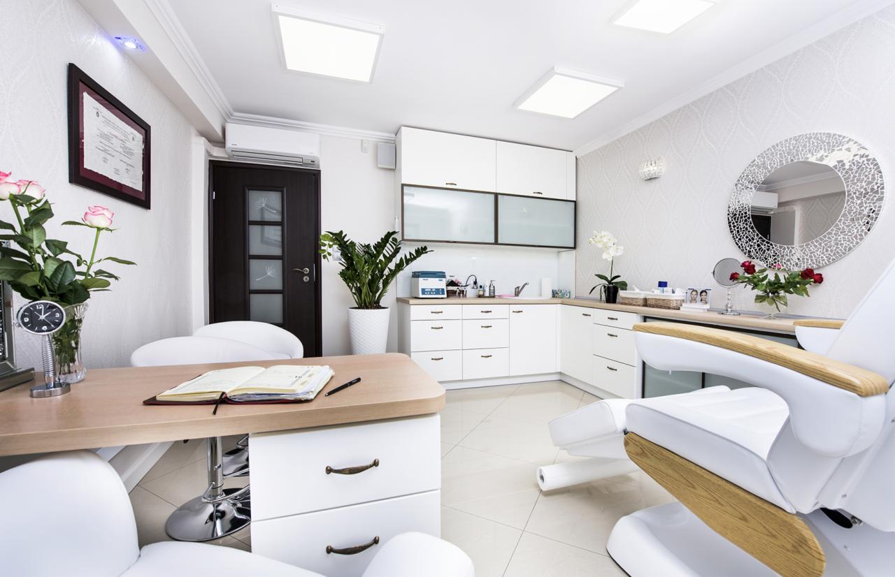 Jasne wnętrze gabinetu kosmetycznego z fotelem zabiegowym, biurkiem z otwartym notatnikiem, kwiatami i dekoracyjnym lustrem na ścianie.
