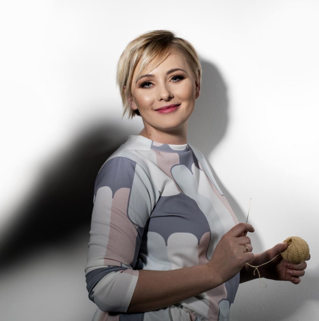 Portret uśmiechniętej kobiety z blond włosami, trzymającej w dłoniach kłębek wełny i szydełko na tle białej ściany z cieniem.