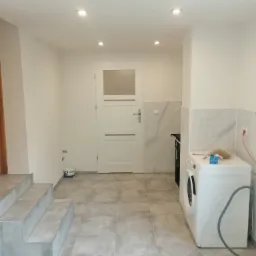 Wykończone wnętrze z szarymi płytkami podłogowymi, schodami i fragmentem białej pralki, białe ściany i drzwi z szybą, widoczne gniazdko elektryczne.