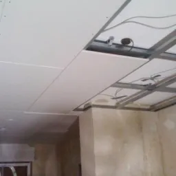 Montaż sufitu podwieszanego z płyt gipsowo-kartonowych na metalowej konstrukcji, widoczne przewody elektryczne i otw&oacute;r na oświetlenie, ściany w stanie surowym.