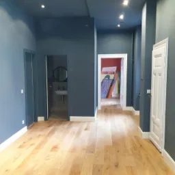 Wnętrze mieszkania po malowaniu ścian na kolor granatowy, z jasną drewnianą podłogą i białymi listwami przypodłogowymi, widoczne drzwi do łazienki z umywalką i lustrem oraz dalsze pomieszczenia...