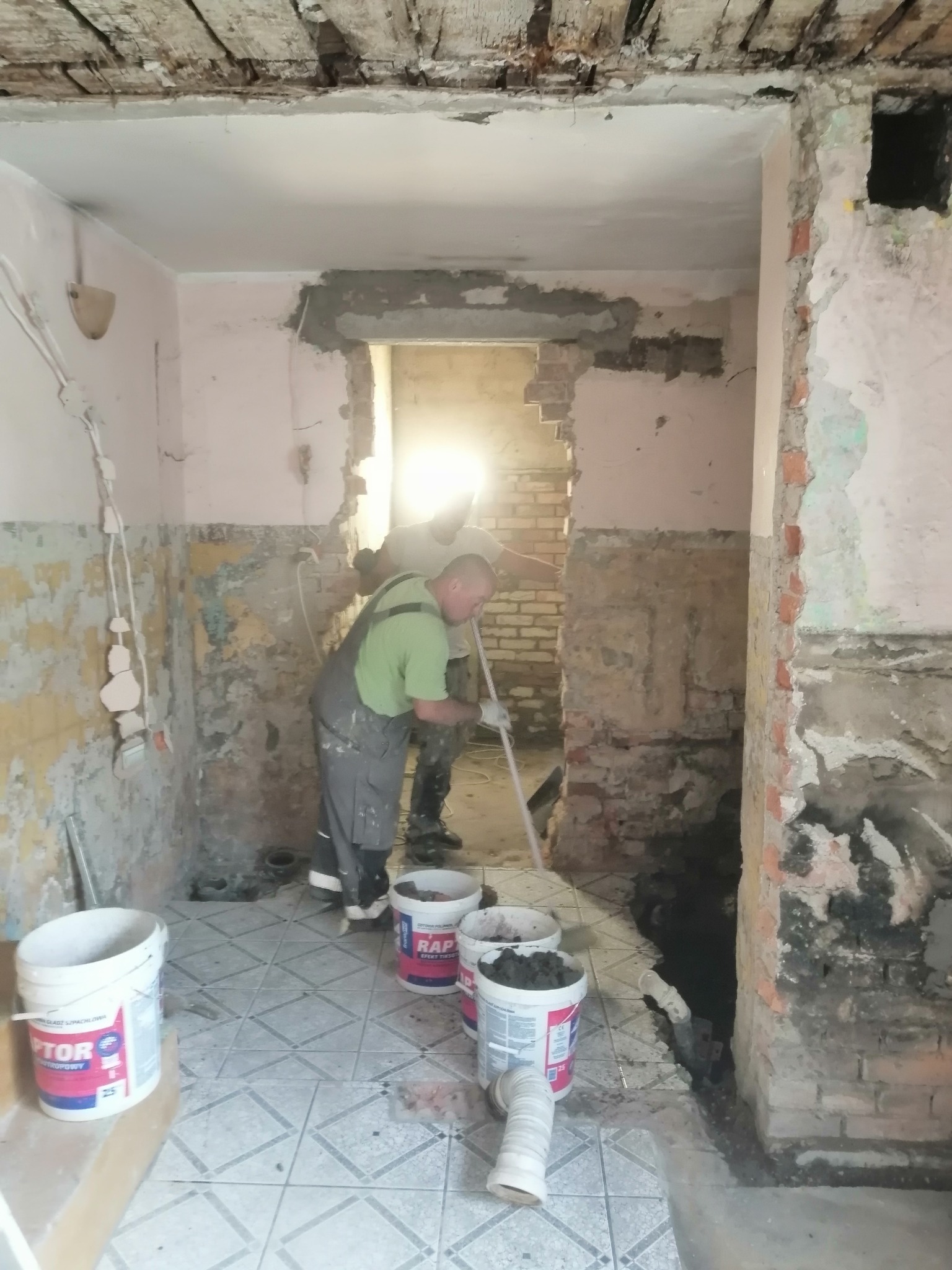 Prace remontowe wewnątrz budynku: przygotowanie ścian pod wykończenie, widoczne wiadra z masą szpachlową i pracownicy w trakcie wyrównywania powierzchni.