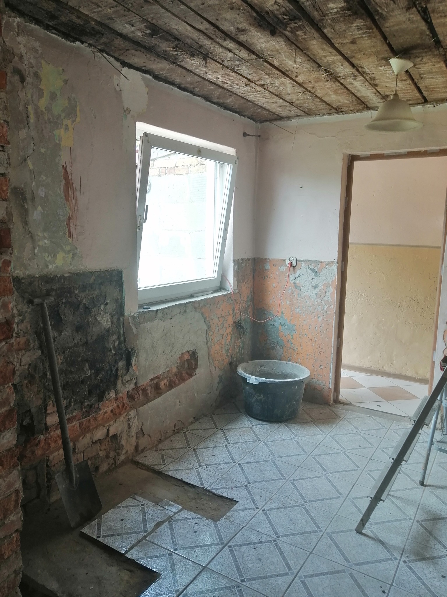 Wnętrze pomieszczenia w trakcie remontu: odsłonięta cegła, częściowo usunięte stare powłoki ścian, widoczne ślady po wcześniejszych warstwach farby, otwarte okno, wiadro i łopata. Zniszczona...