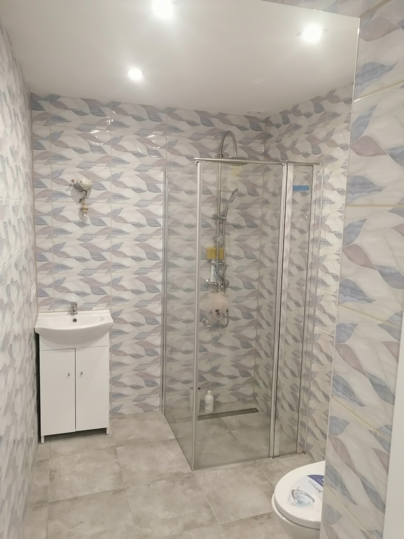 Wykończona łazienka z kabiną prysznicową, umywalką z szafką i toaletą. Na ścianach i podłodze płytki w odcieniach szarości i bieli z motywem liści. Widoczna niedokończona instalacja oświetleniowa.